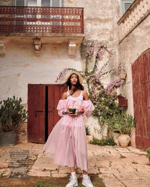 A young woman in a pink dress and straw hat poses in front of an aged stone building with bougainvillea and rustic red doors.