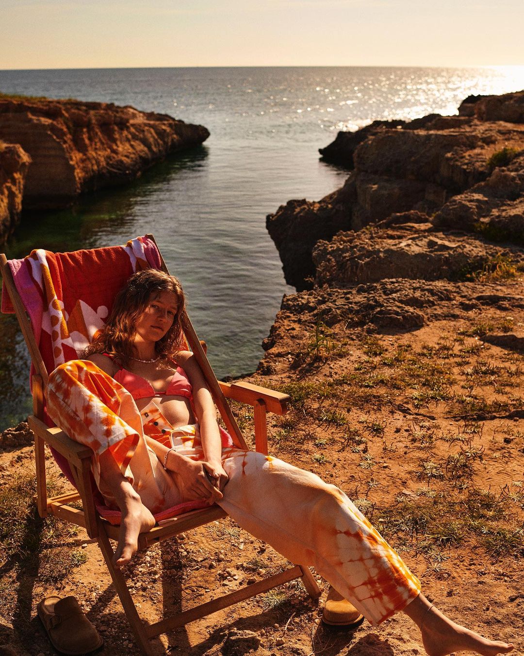 Adult woman in tie-dye pants and bikini top reclines in a folding chair by the ocean at sunset, with orange rocks and shoreline.