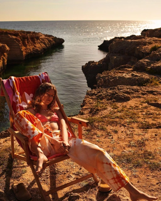 Adult woman in tie-dye pants and bikini top reclines in a folding chair by the ocean at sunset, with orange rocks and shoreline.