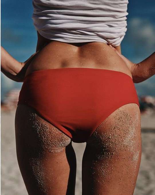 Woman wearing red bikini bottoms, rear view, standing on a beach with sand visible on her skin and a blue sky in the background.