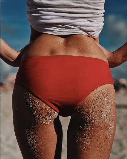 Woman wearing red bikini bottoms, rear view, standing on a beach with sand visible on her skin and a blue sky in the background.