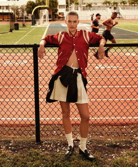 A young adult woman poses on an athletic field in a fashion editorial shot, wearing a letterman jacket, pleated skirt, socks, and loafers.