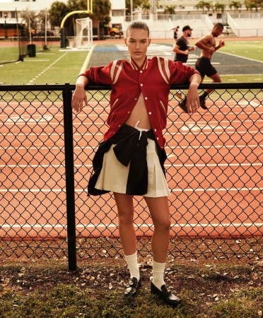 A young adult woman poses on an athletic field in a fashion editorial shot, wearing a letterman jacket, pleated skirt, socks, and loafers.