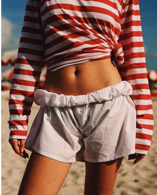 A slender woman poses outdoors, showcasing a red and white striped top, white shorts, and tanned skin, with a beach backdrop.