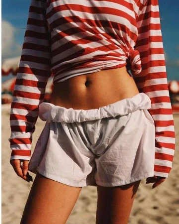 A slender woman poses outdoors, showcasing a red and white striped top, white shorts, and tanned skin, with a beach backdrop.