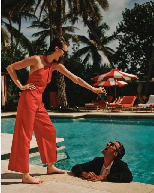 Woman pours champagne on a man in a pool on a sunny day; she wears a red jumpsuit, and he wears a dark suit jacket and sunglasses.