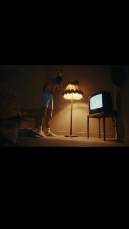 An indoor low-angle shot reveals a person in pajamas holding a doll next to a lamp and old television in a dimly lit room at night.