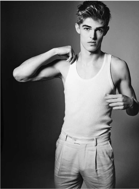 Black-and-white portrait of a young adult male with short hair, wearing a white ribbed tank top and light-colored pants, posing indoors.