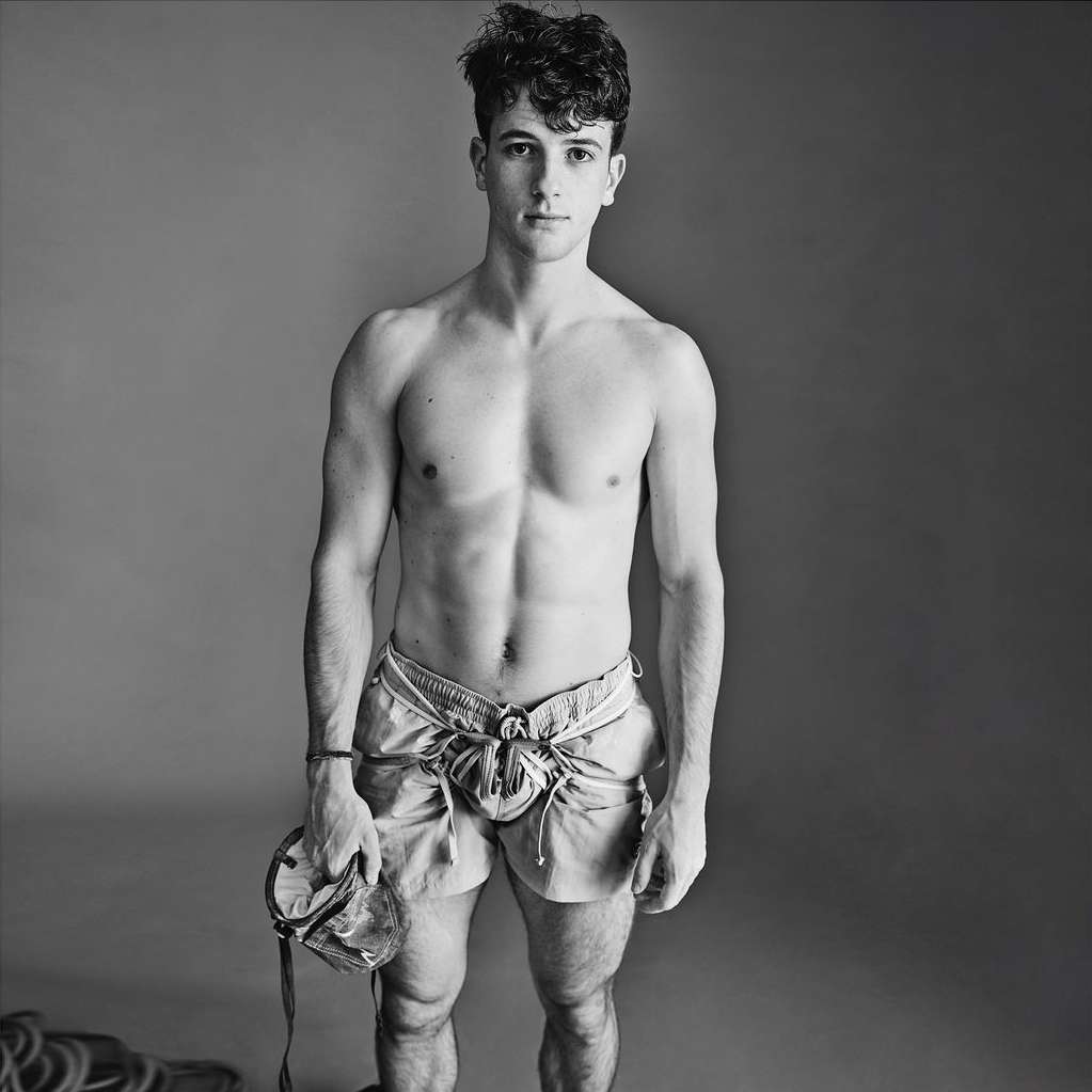 Monochrome portrait of a muscular shirtless man in shorts holding climbing equipment, posing against a gray background in a studio.