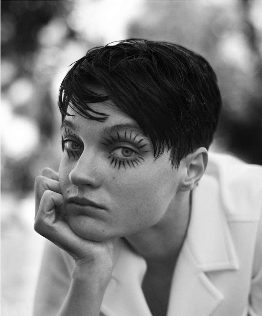 Portrait of a stylish short-haired woman with dramatic black and white eye makeup, gazing forward with neutral expression, outdoors.