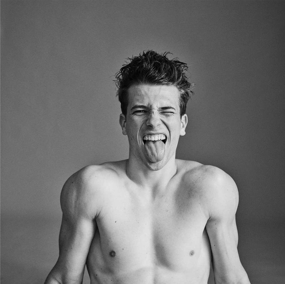 A black and white portrait of a shirtless young man with dark hair sticking his tongue out and squinting against a grey background.