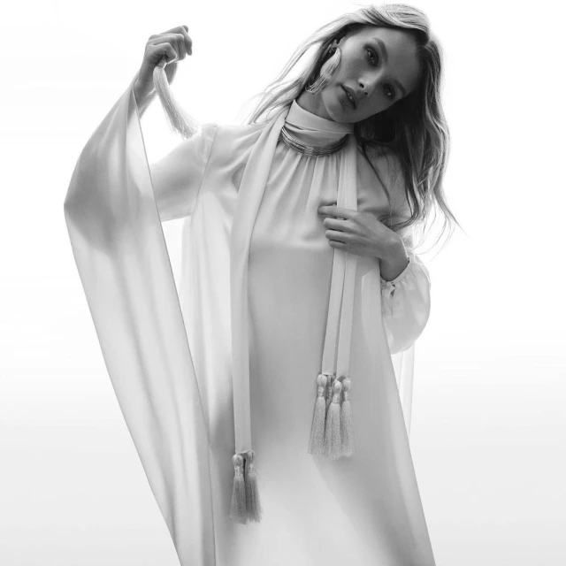 Black and white studio shot of a blonde woman in an elegant white gown with draped sleeves, posing with a tasseled sash.