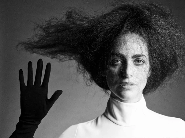 Monochrome portrait of a woman with voluminous dark hair and a gloved hand raised against a grey background.