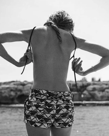 Monochrome rear view of a woman adjusting patterned shorts straps outdoors near water, photographed by Giampaolo Sgura.