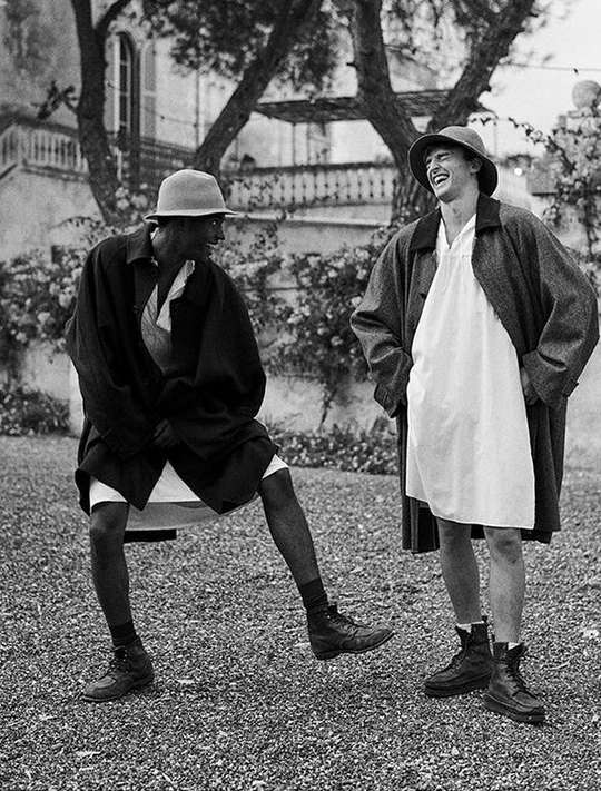 Two men laughing outdoors in loose outerwear and hats against a building and foliage background, photographed in black and white.