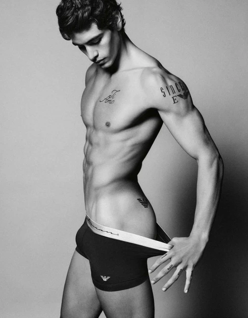 Black and white portrait of a fit, shirtless man in briefs holding the waistband with tattoos posing indoors against a grey background.