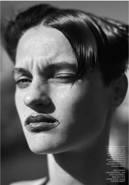 Monochrome portrait of a young adult woman with a short haircut and dark lipstick, set against a blurred outdoor background.
