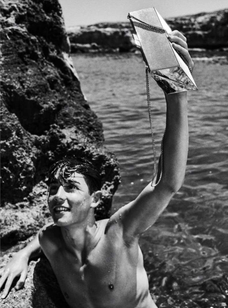 Teenage boy smiles while holding a shiny purse in front of ocean water and a rocky shoreline during a sunny day. Black and white.