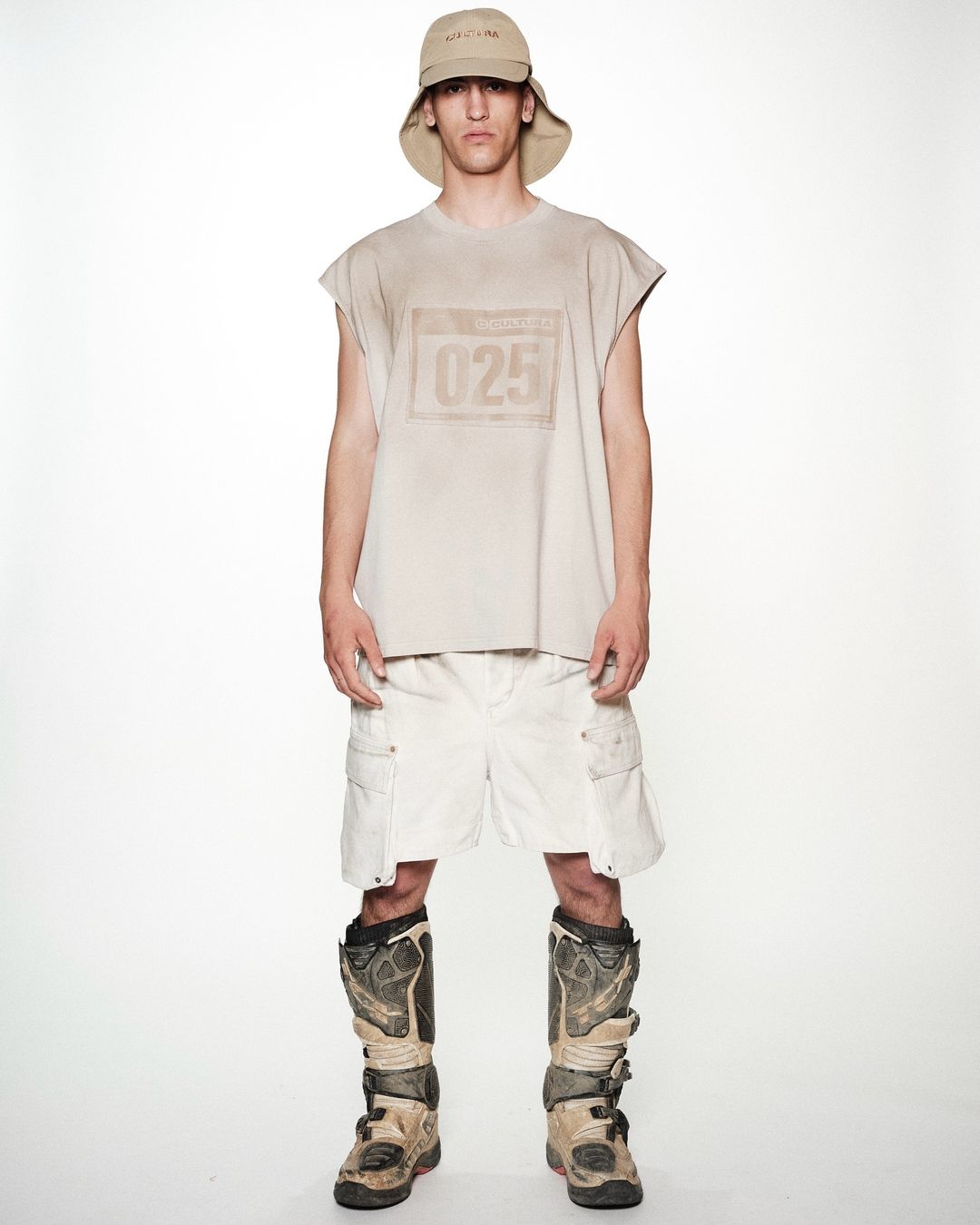 Full shot of a young adult man in a sleeveless tan shirt, white shorts, a bucket hat, and tan motocross boots, standing against a white backdrop.