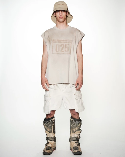 Full shot of a young adult man in a sleeveless tan shirt, white shorts, a bucket hat, and tan motocross boots, standing against a white backdrop.