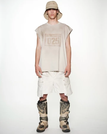 Full shot of a young adult man in a sleeveless tan shirt, white shorts, a bucket hat, and tan motocross boots, standing against a white backdrop.