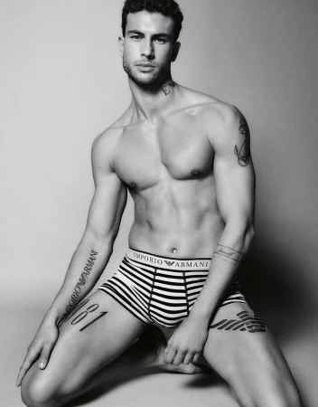 Black and white portrait of a shirtless male model wearing branded striped briefs against a seamless studio background.