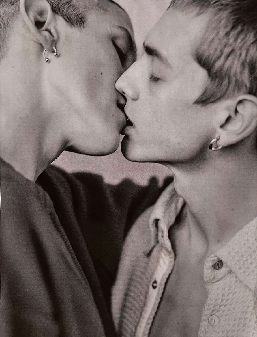 Close-up of two adult men kissing, with visible earrings and collared shirts, set against a blurred, neutral background in a monochrome photograph.