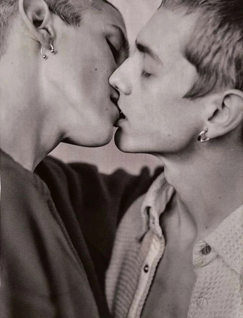Close-up of two adult men kissing, with visible earrings and collared shirts, set against a blurred, neutral background in a monochrome photograph.
