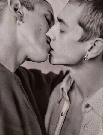Close-up of two adult men kissing, with visible earrings and collared shirts, set against a blurred, neutral background in a monochrome photograph.