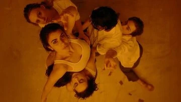 Overhead shot of five people lying on a reflective surface; dark-haired woman in a white top is a focal point, warm lighting.