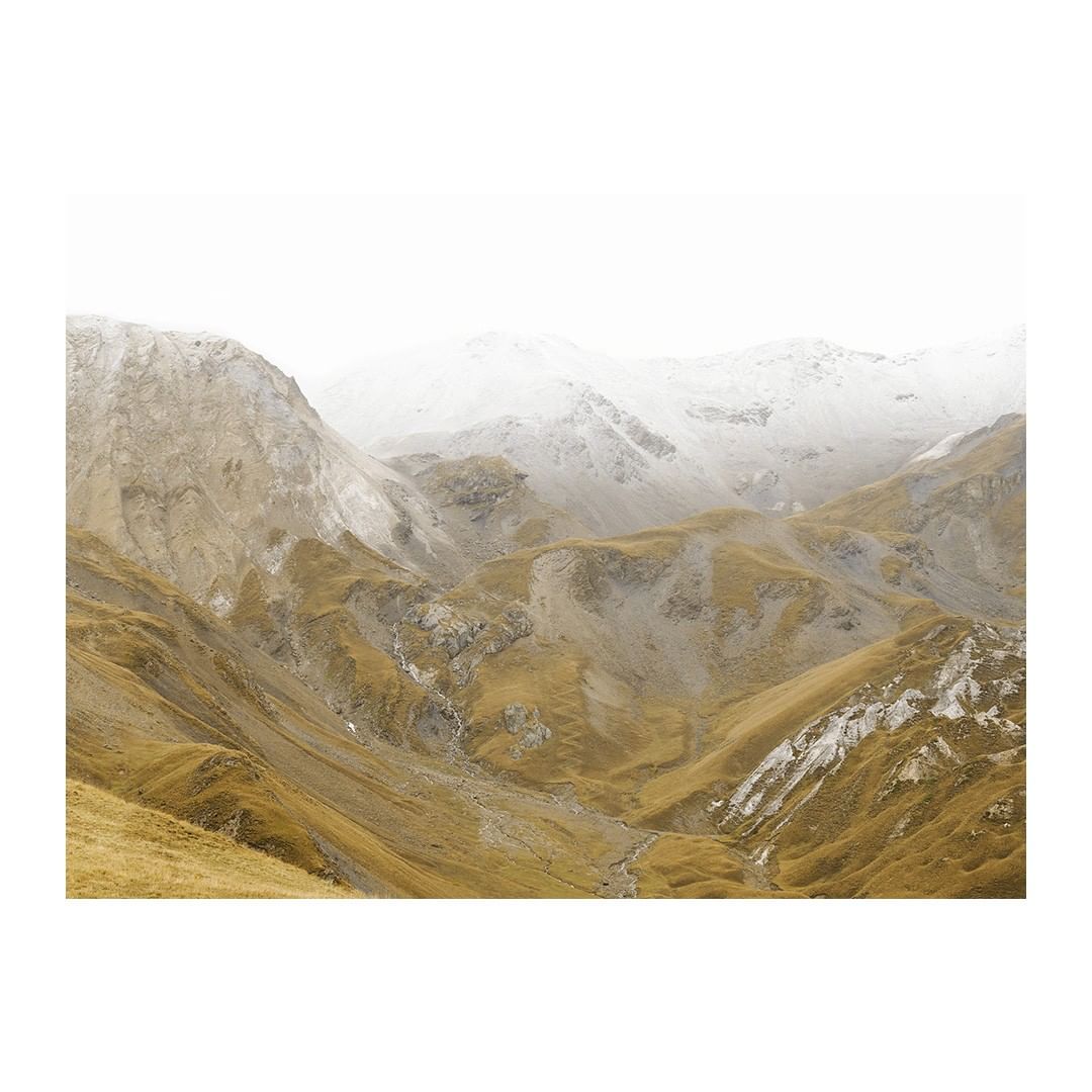 Scenic view of a vast mountain range with golden grasses, rocky terrain, and snow-capped peaks under a bright, hazy sky.