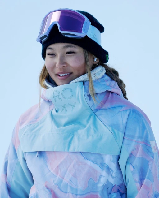 Close-up portrait of a smiling young Asian woman in a snow hat and goggles, wearing a multicolored Roxy brand jacket in an outdoor setting.
