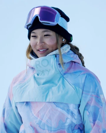 Close-up portrait of a smiling young Asian woman in a snow hat and goggles, wearing a multicolored Roxy brand jacket in an outdoor setting.