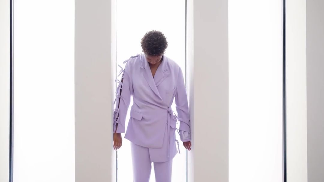 Adult female poses in a pastel purple jacket and trousers in front of a window with bright light, creating a fashion-forward editorial image.
