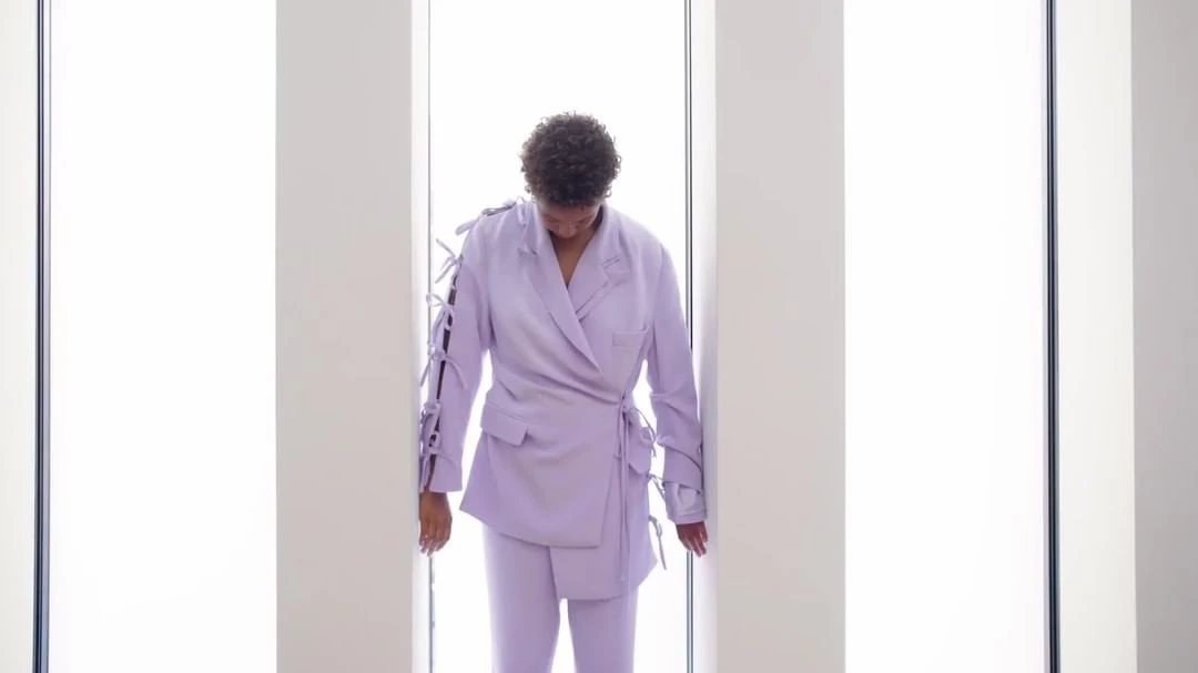 Adult female poses in a pastel purple jacket and trousers in front of a window with bright light, creating a fashion-forward editorial image.