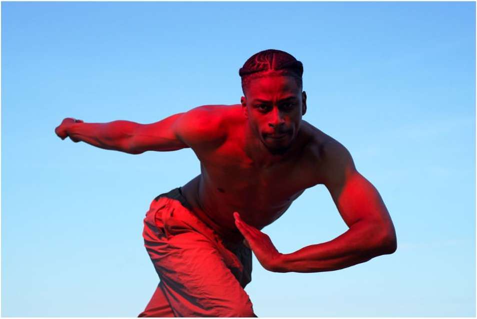 Athletic man in dynamic pose, shirtless in grey pants, strongly lit in red against a clear blue sky, full frame, medium shot.