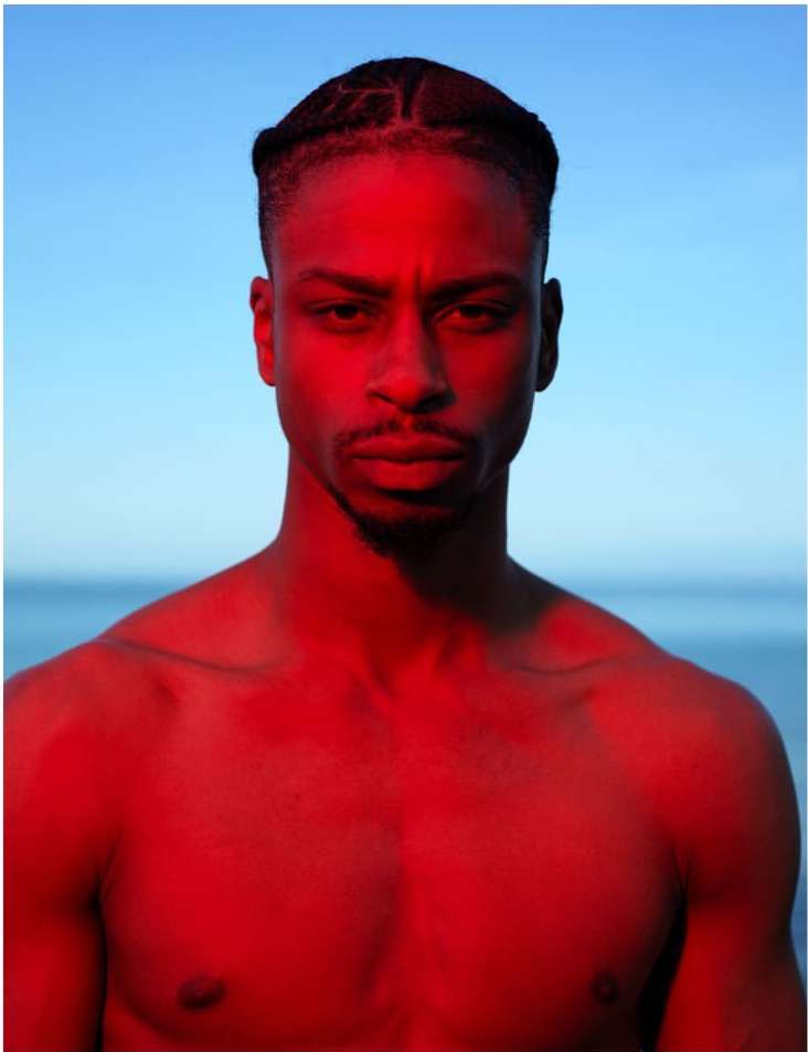 Portrait of a black man with braided hair under intense red lighting against a blue sky and horizon