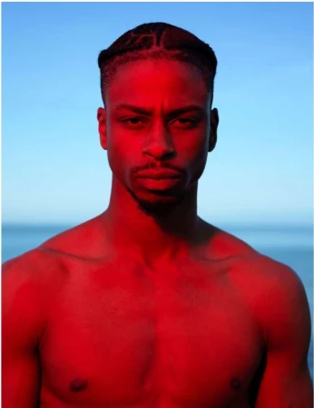 Portrait of a black man with braided hair under intense red lighting against a blue sky and horizon
