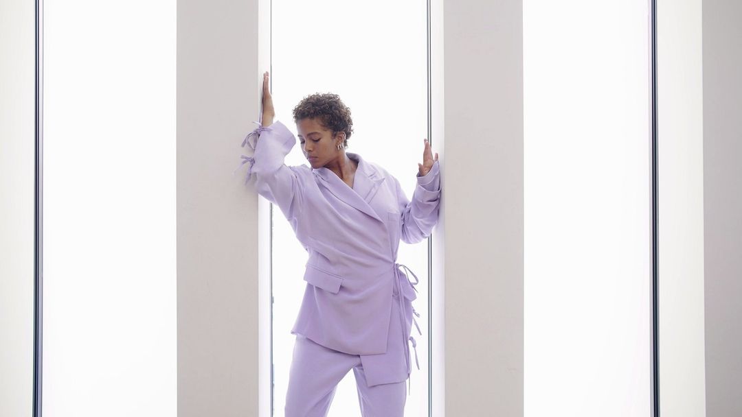 Woman in lilac pantsuit poses between bright light panels, fashion portrait, studio shot with indoor setting, minimalist fashion.