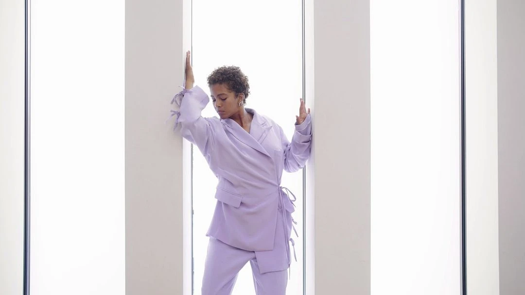 Woman in lilac pantsuit poses between bright light panels, fashion portrait, studio shot with indoor setting, minimalist fashion.