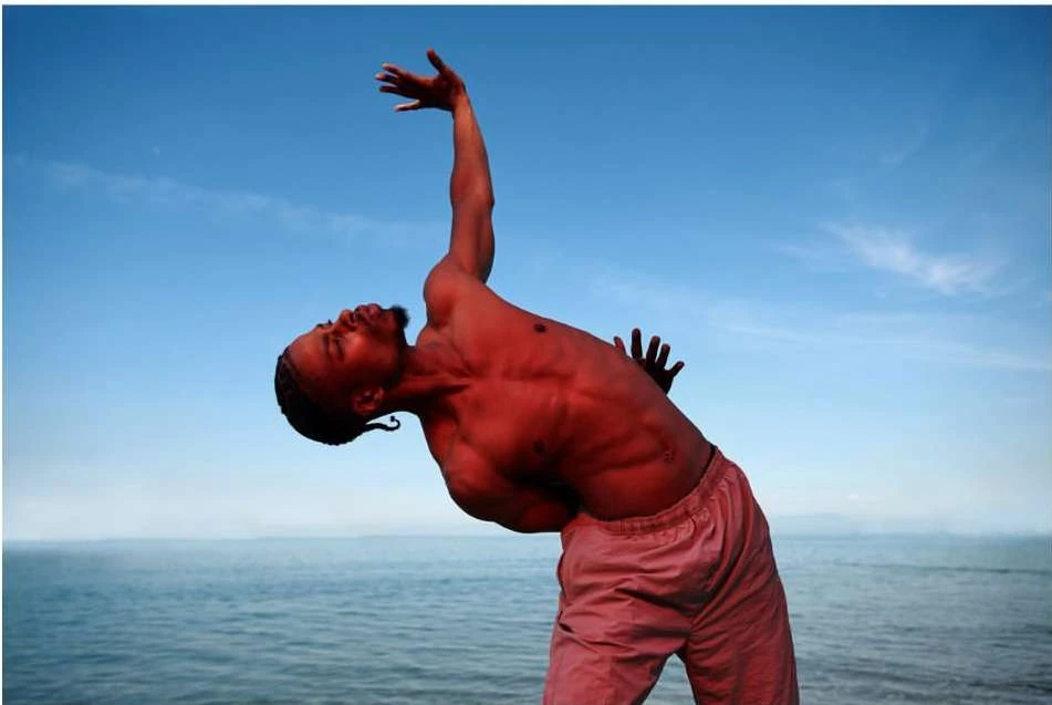 Man with athletic build poses with arms raised toward the bright blue sky, with the ocean in the distance, wearing pink shorts.