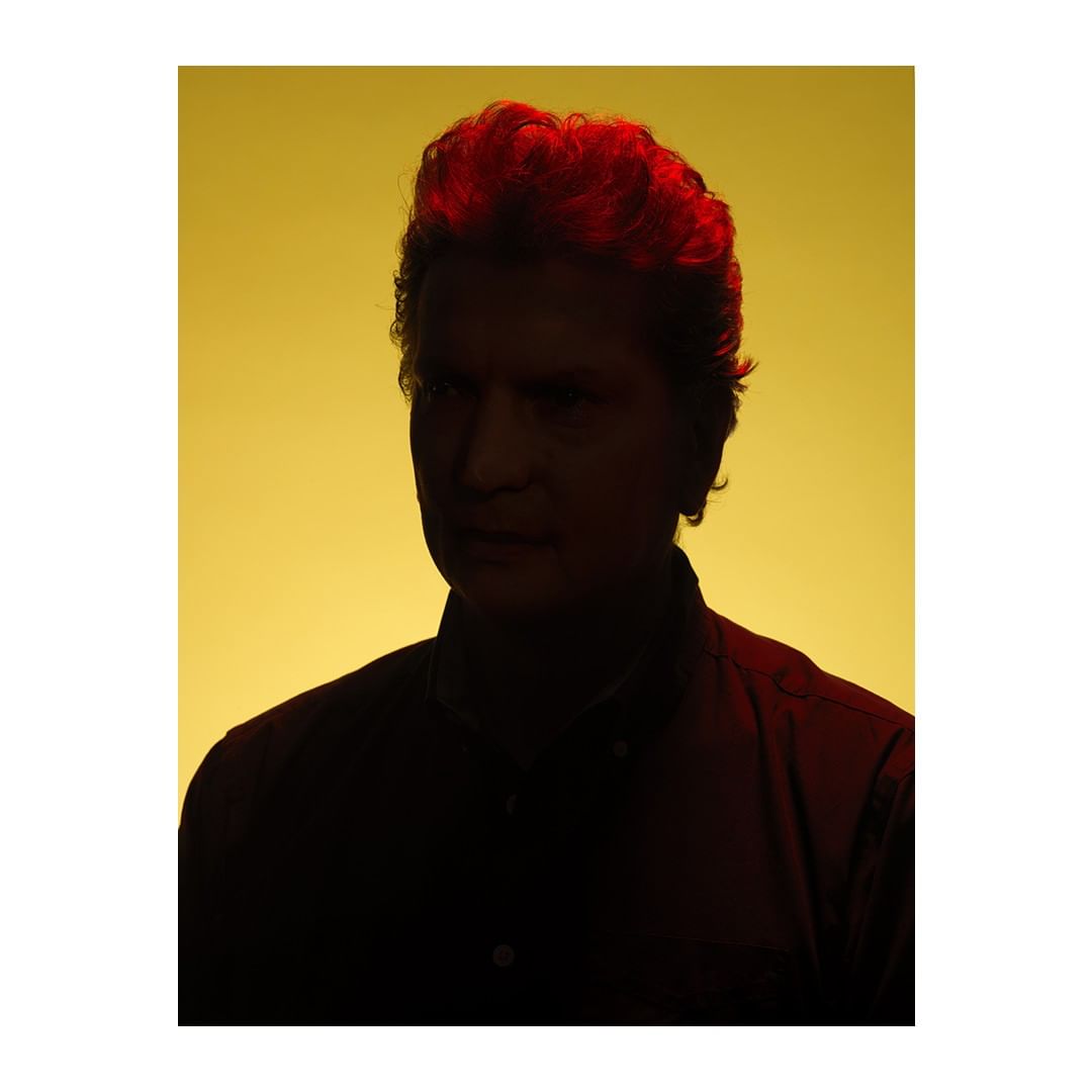 Portrait of a silhouetted adult male with auburn hair against a yellow background in a studio setting. The subject is looking to the left.