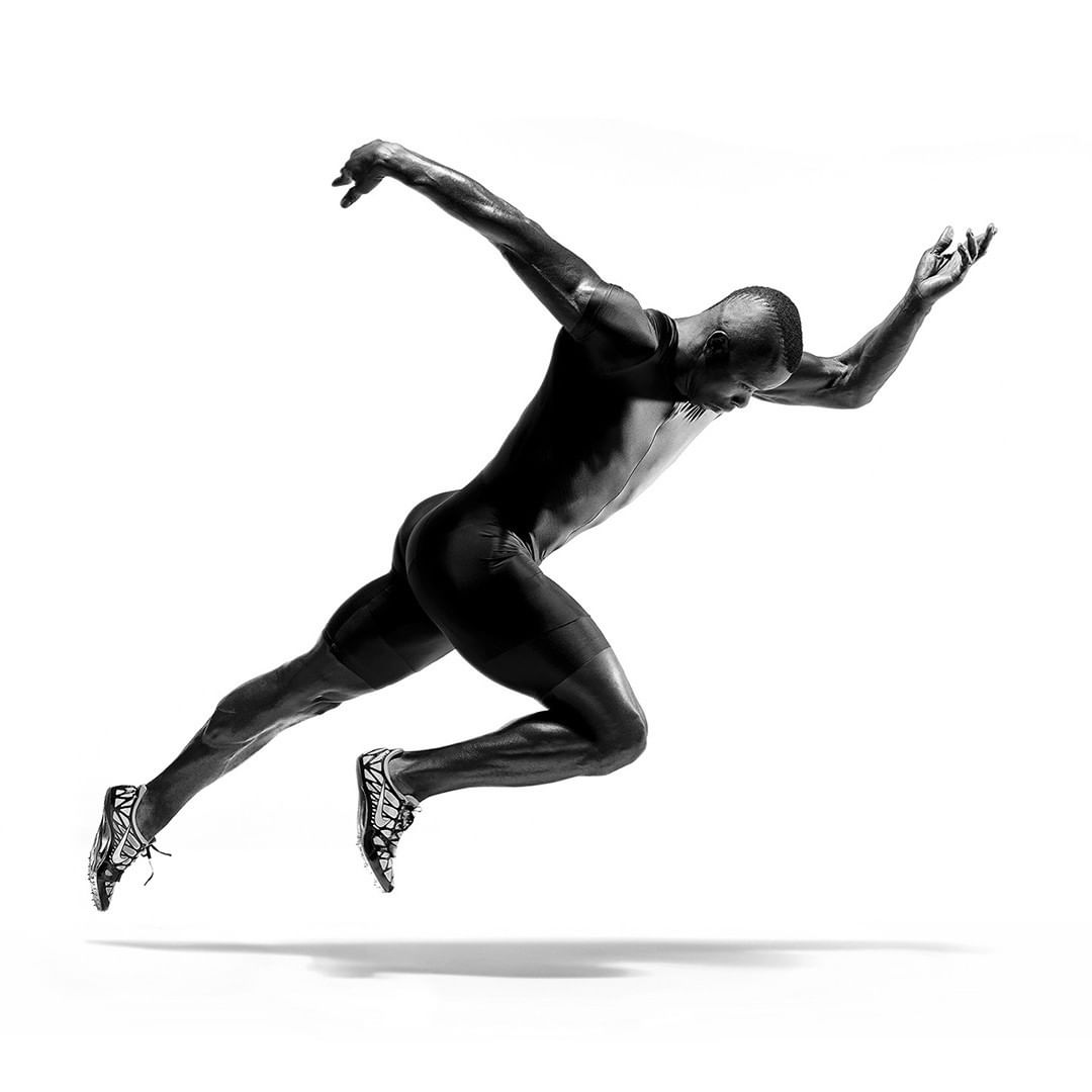 Monochrome studio shot of a Black male athlete in mid-run, muscular build, wearing athletic gear, captured against a stark white background.