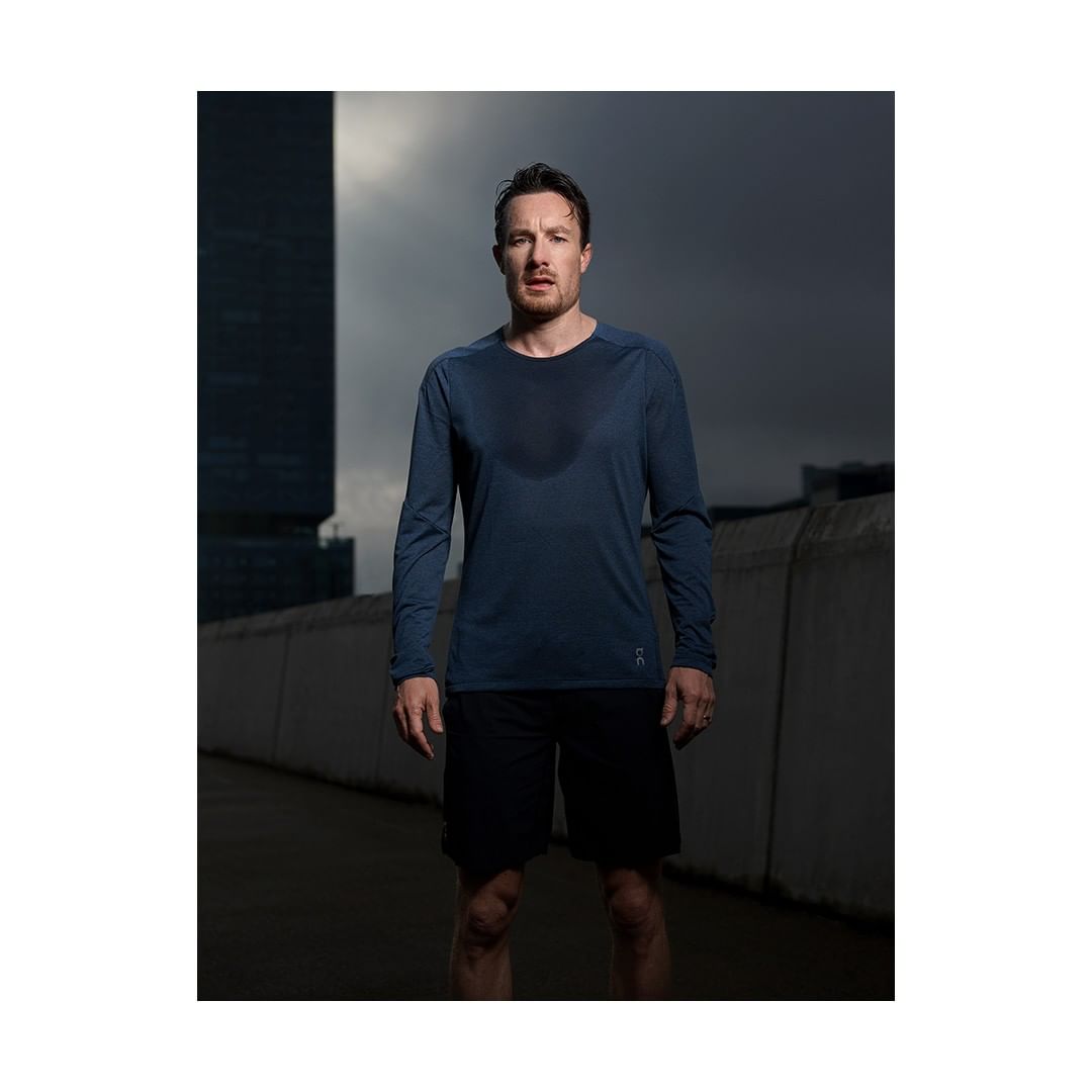 Athletic man in long-sleeve shirt and shorts stands on urban street in front of tall buildings, showcasing fitness wear.