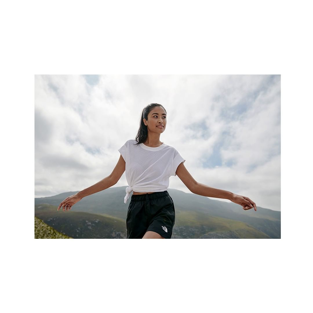 An adult woman with arms outstretched wearing athletic shorts and a white T-shirt stands outdoors with a mountain range background.