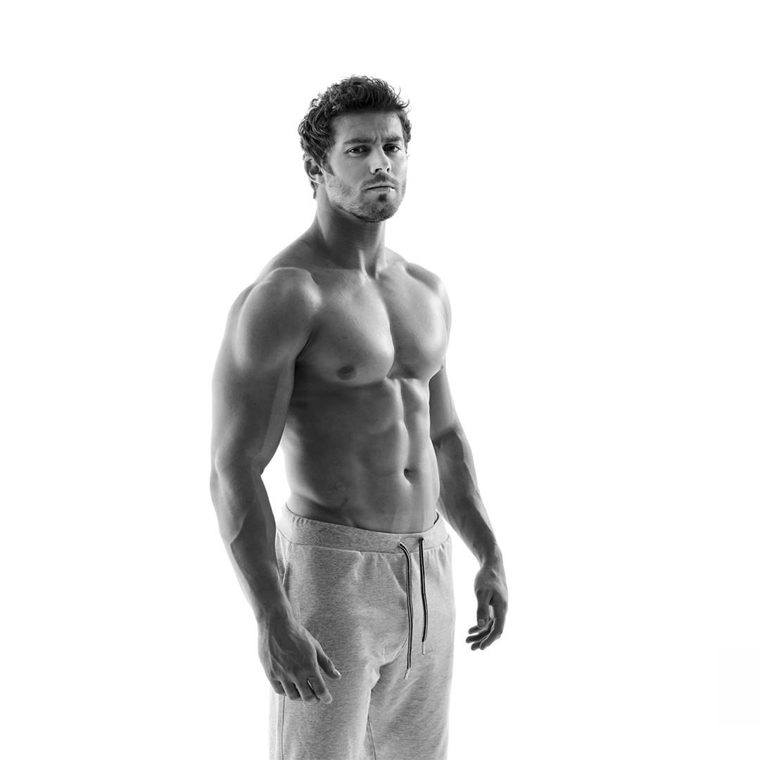 Black and white studio shot of a muscular adult male with a serious expression, standing in gray sweatpants against a white background.