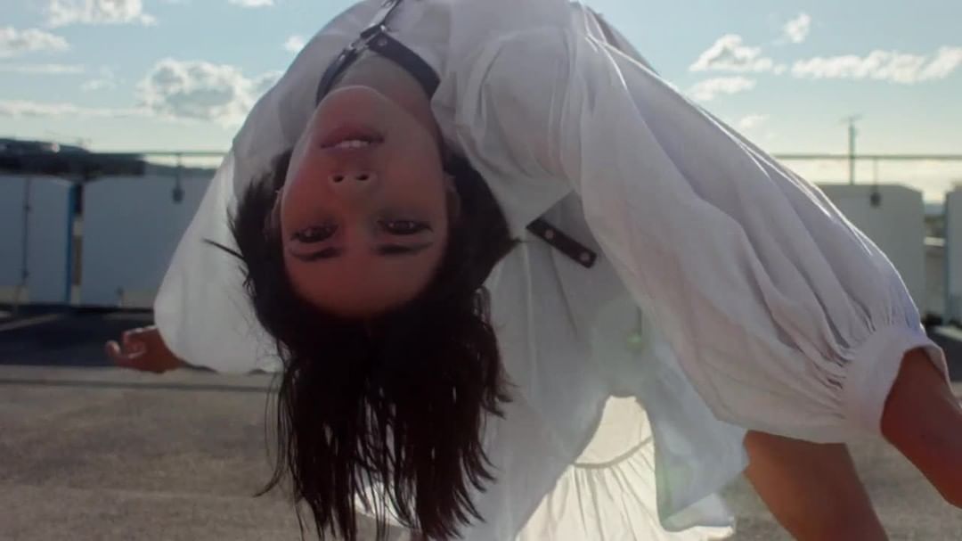 Brunette woman in white shirt with black harness poses upside down on rooftop with city skyline and cloudy sky in the background.