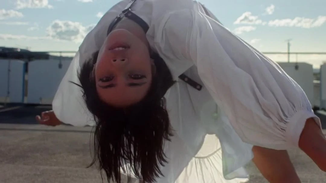 Brunette woman in white shirt with black harness poses upside down on rooftop with city skyline and cloudy sky in the background.