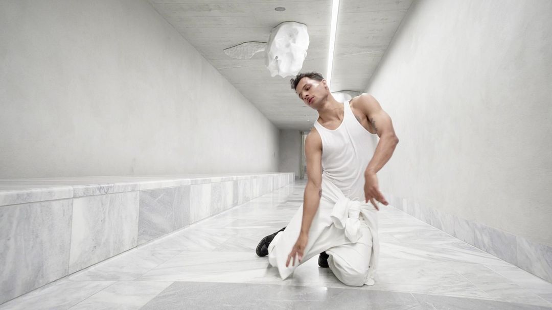 Adult male in white tank top and trousers kneeling in modern corridor with white walls and marble floor; sculpture on ceiling.