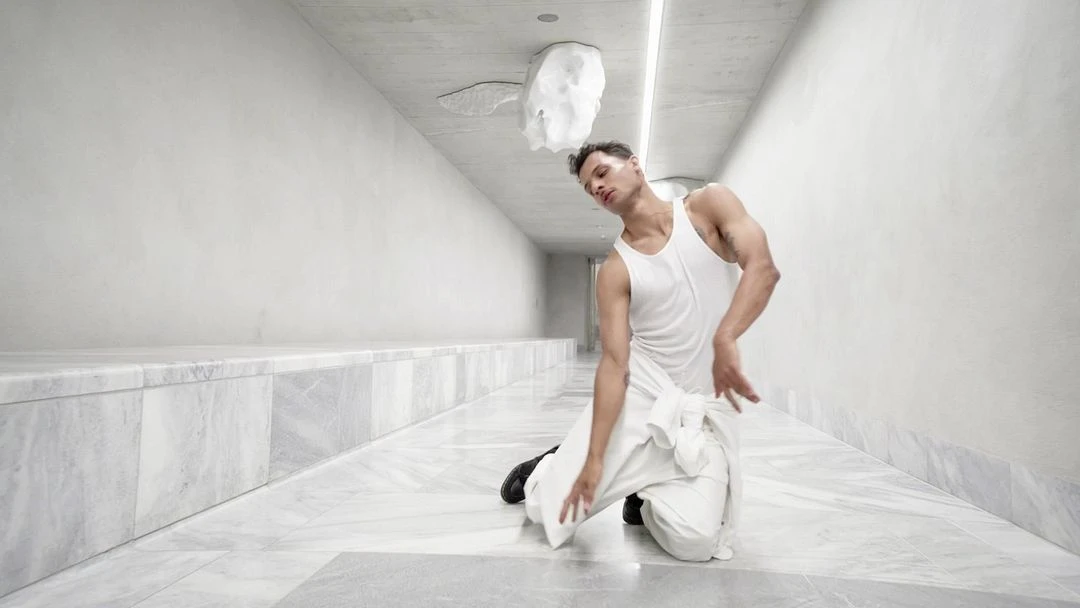 Adult male in white tank top and trousers kneeling in modern corridor with white walls and marble floor; sculpture on ceiling.